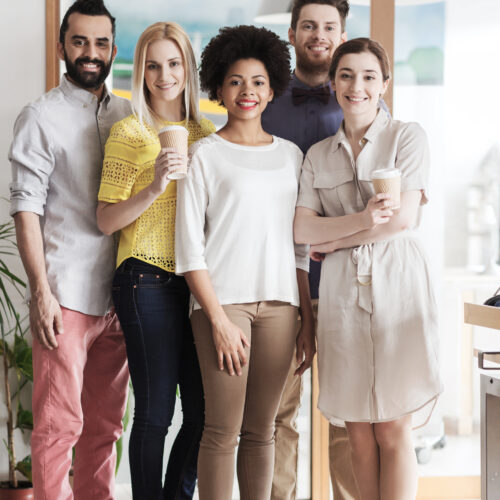 business, startup, people, drinks and teamwork concept - happy smiling international creative team with disposable paper coffee cups in office